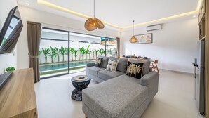A modern living room with a grey sofa, a coffee table, and a TV. There is a large window with a view of a pool and greenery outside. - Brand New Beautiful Stylish 2 BR Villa in Canggu (Bali)