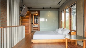 A modern bedroom with a wooden floor, a large bed, a wooden bedside table, and a bathroom with a glass shower enclosure. - SMARANALA  (Nusa Dua)
