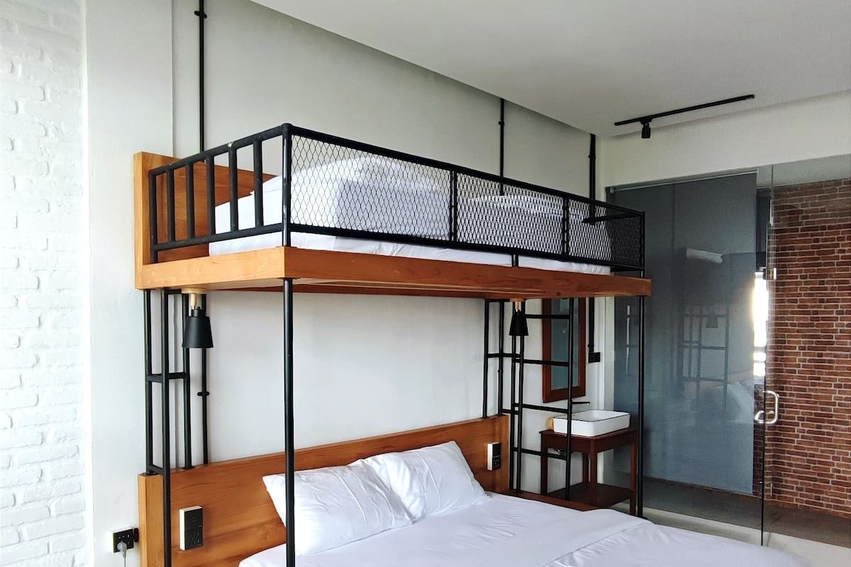 A bunk bed with a wooden frame and white bedding. There is a desk and chair to the right of the bed. The room has white walls and a tiled floor.