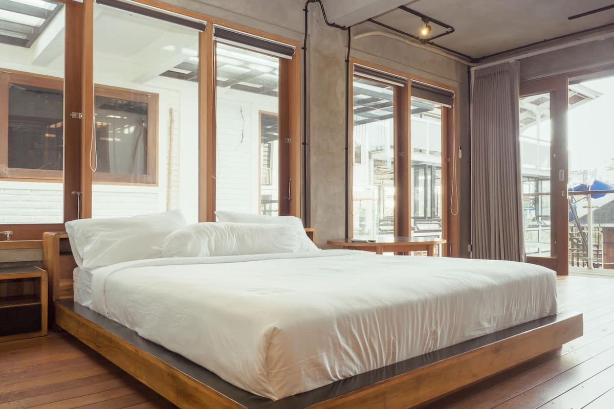 A large bed in a room with wooden flooring, large windows, and a view of the outside.