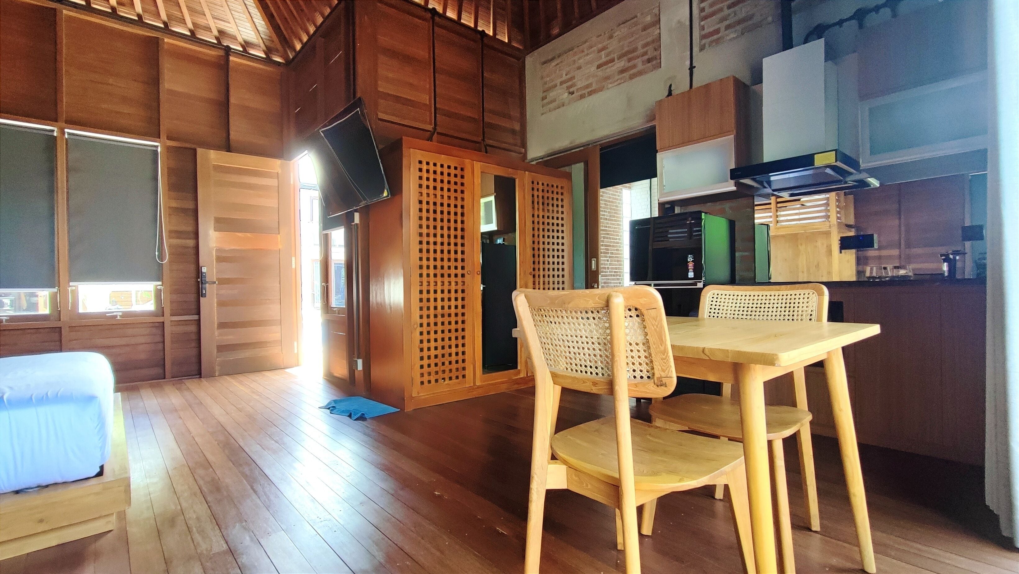 A wooden-floored room with a bed, a dining table with chairs, a kitchen area with a refrigerator, and a TV mounted on the wall.