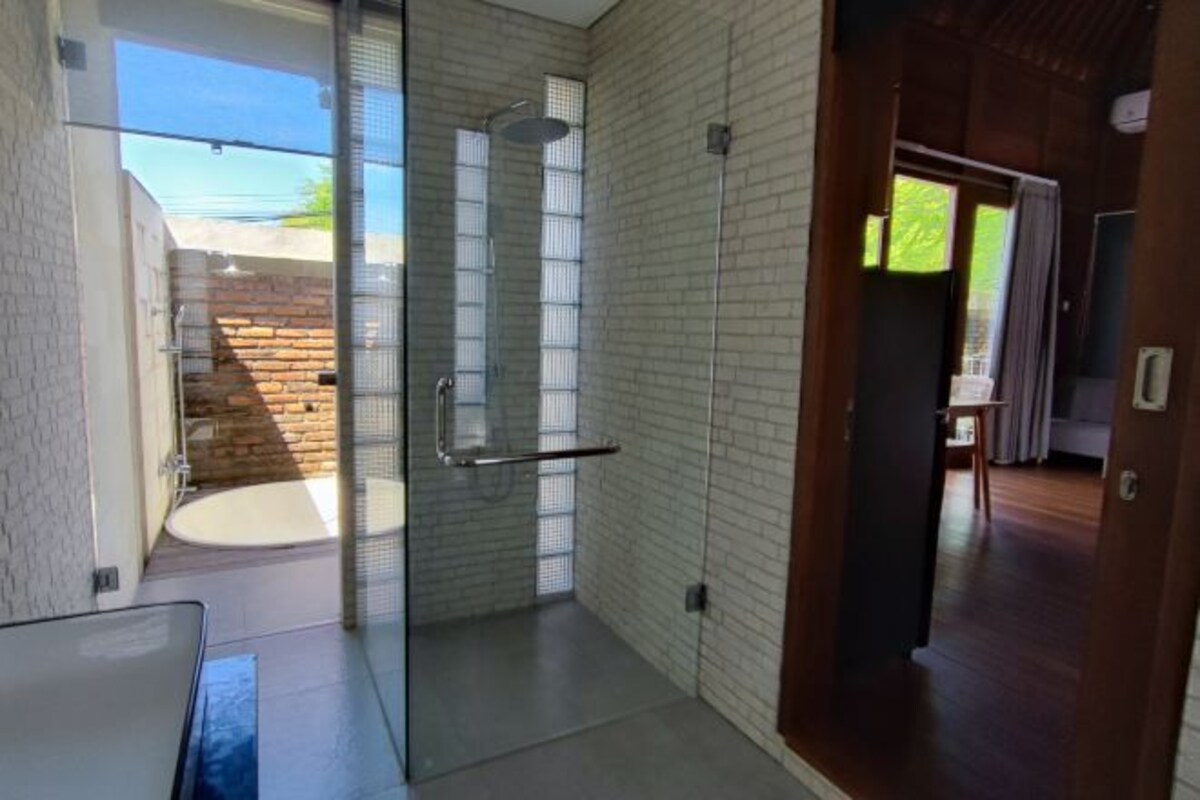 A modern bathroom with a glass shower enclosure, a visible outdoor area with a brick wall, and a wooden floor.