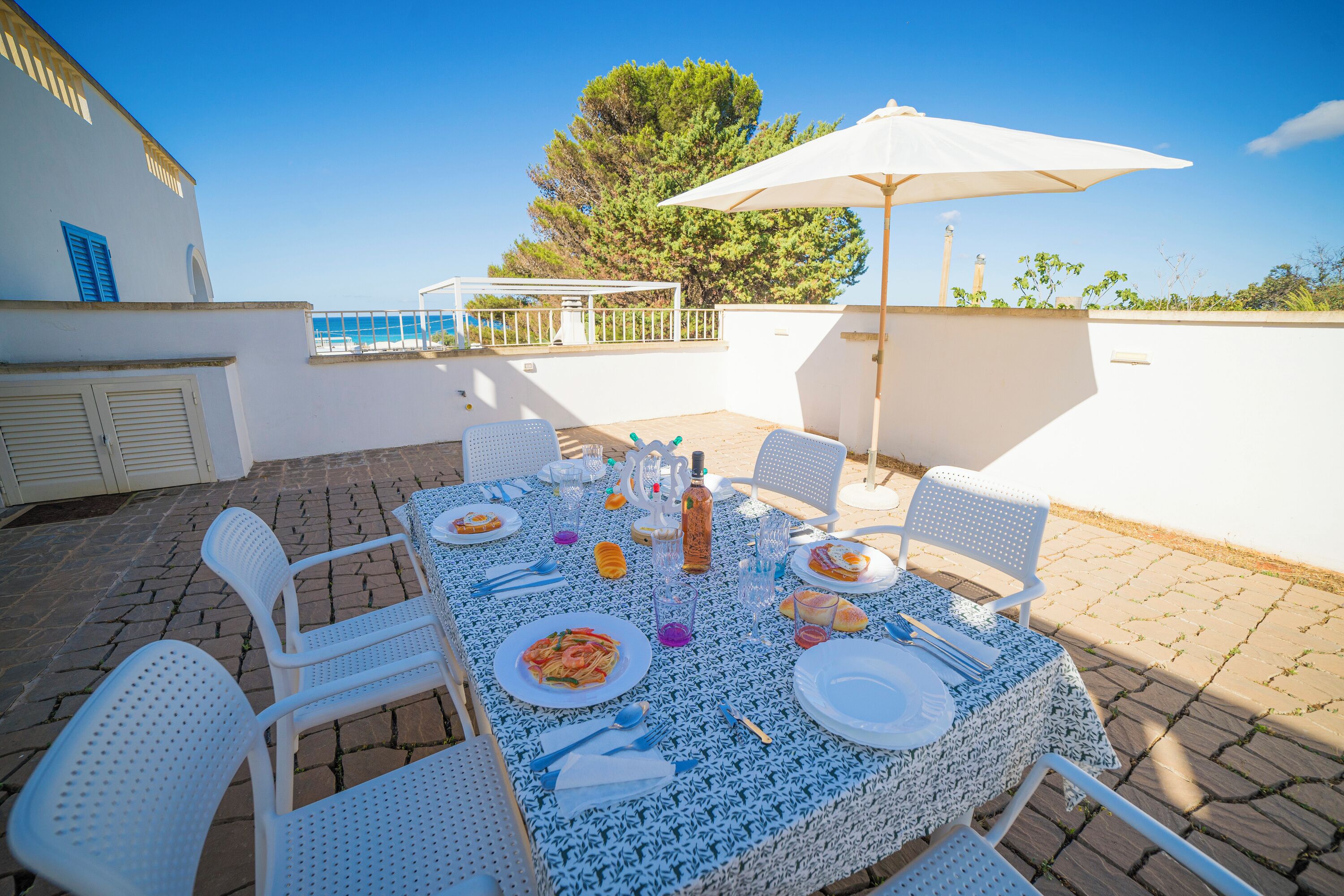 An outdoor patio with a table set for dining, surrounded by white chairs, and an umbrella providing shade.