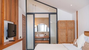 A modern bedroom with a large bed, a television on a wooden shelf, a bathroom with a sink and mirror, and a wardrobe with a woven design. - Mija Bhuana Agung Villa by Betterplace (Canggu)