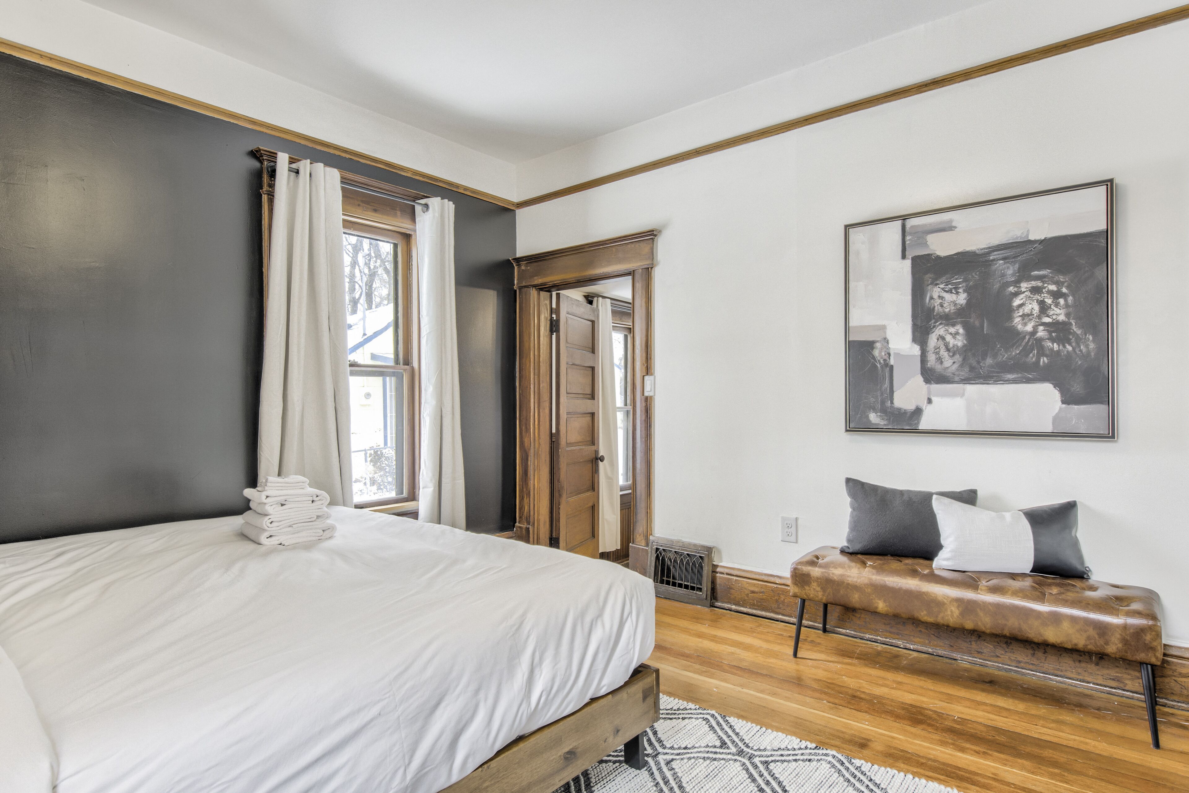 Un dormitorio con una cama, un banco, una obra de arte enmarcada y una puerta que da a otra habitación.