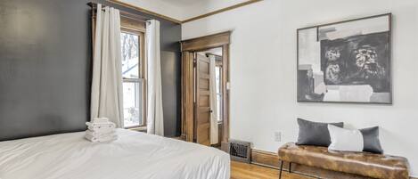 Un dormitorio con una cama, un banco, una obra de arte enmarcada y una puerta que da a otra habitación.
