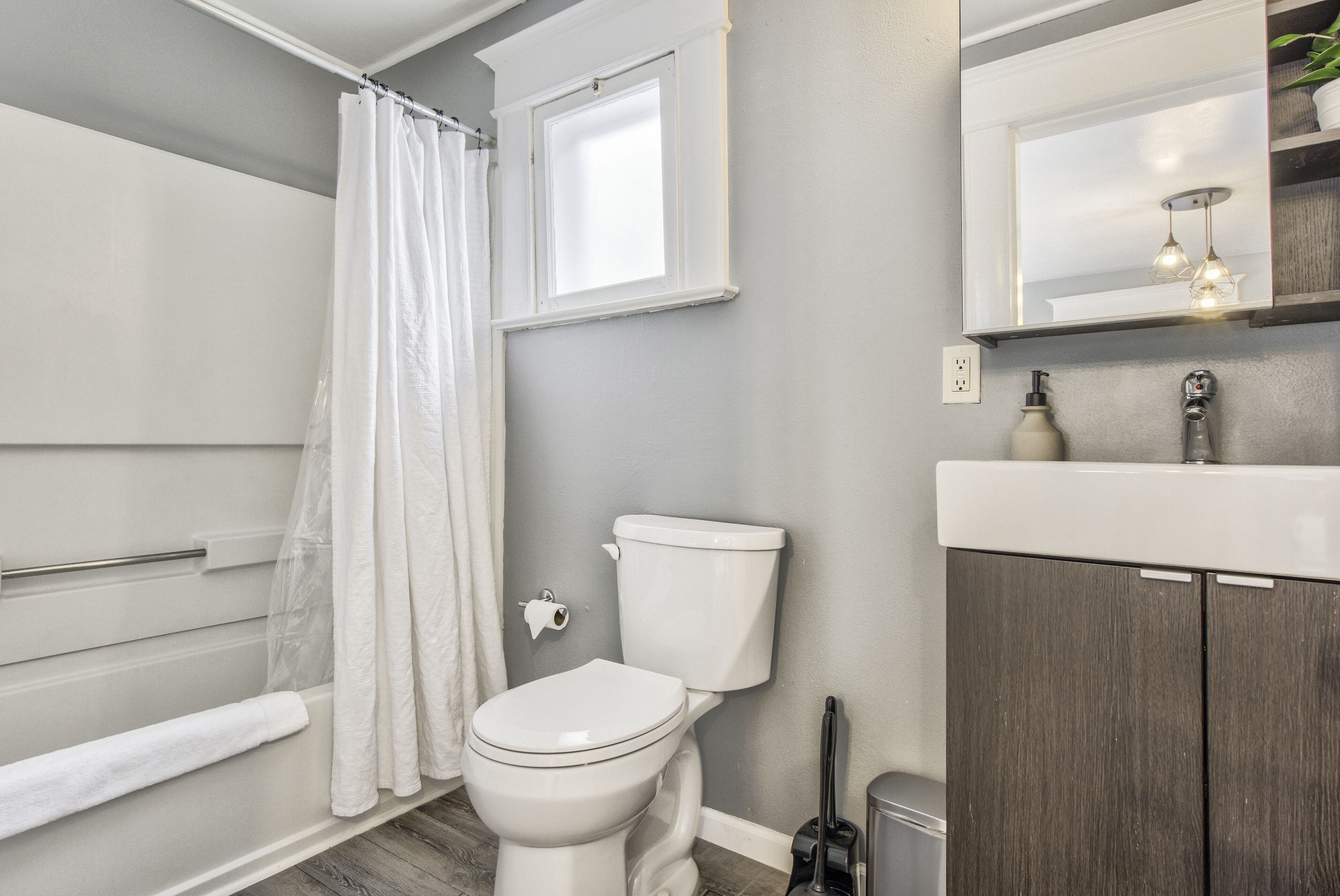 Ein Badezimmer mit Toilette, Duschvorhang, Fenster, Spiegel und Waschbecken mit Schrank.