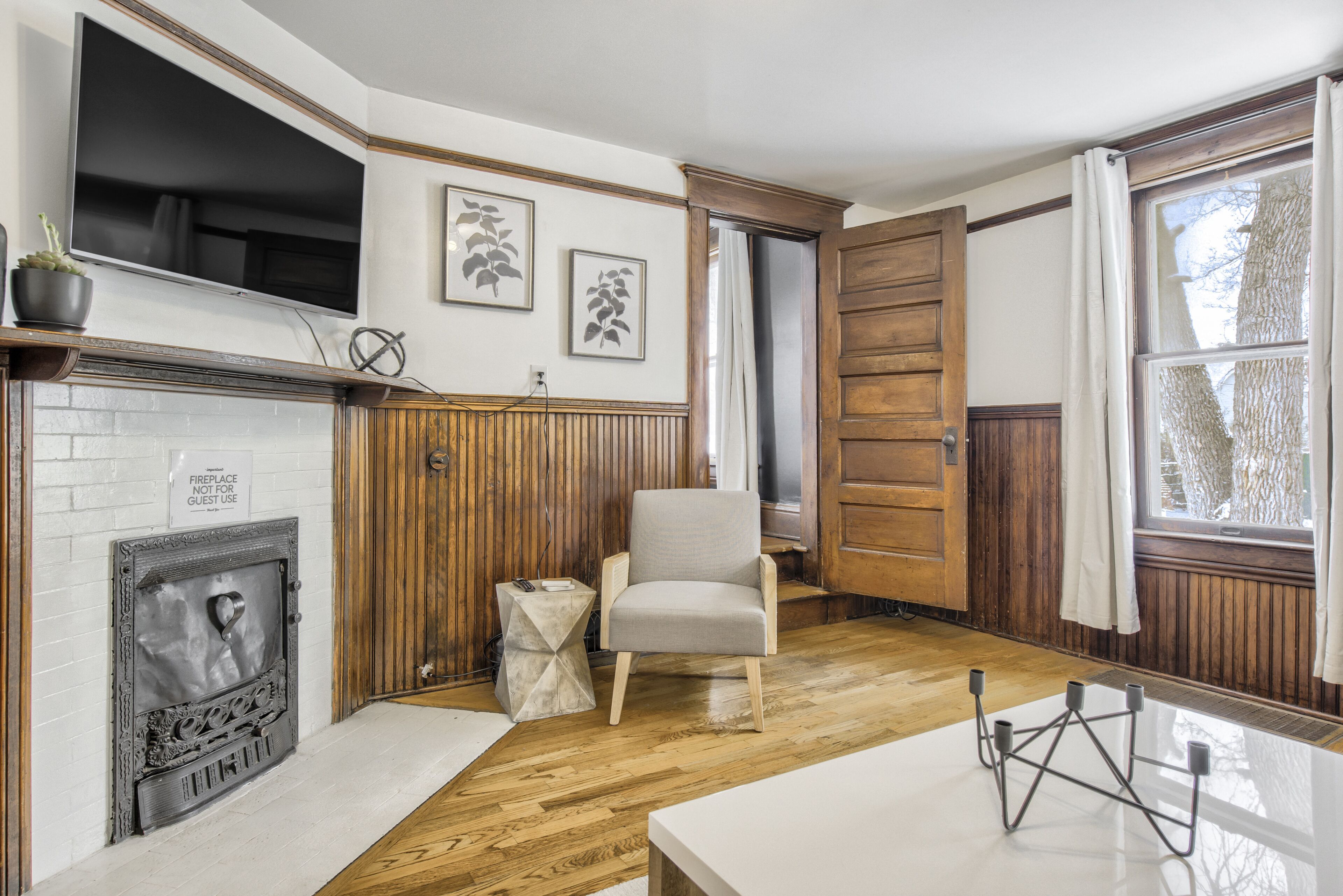 A room with a fireplace, wooden paneling, a white chair, and a large window with curtains.