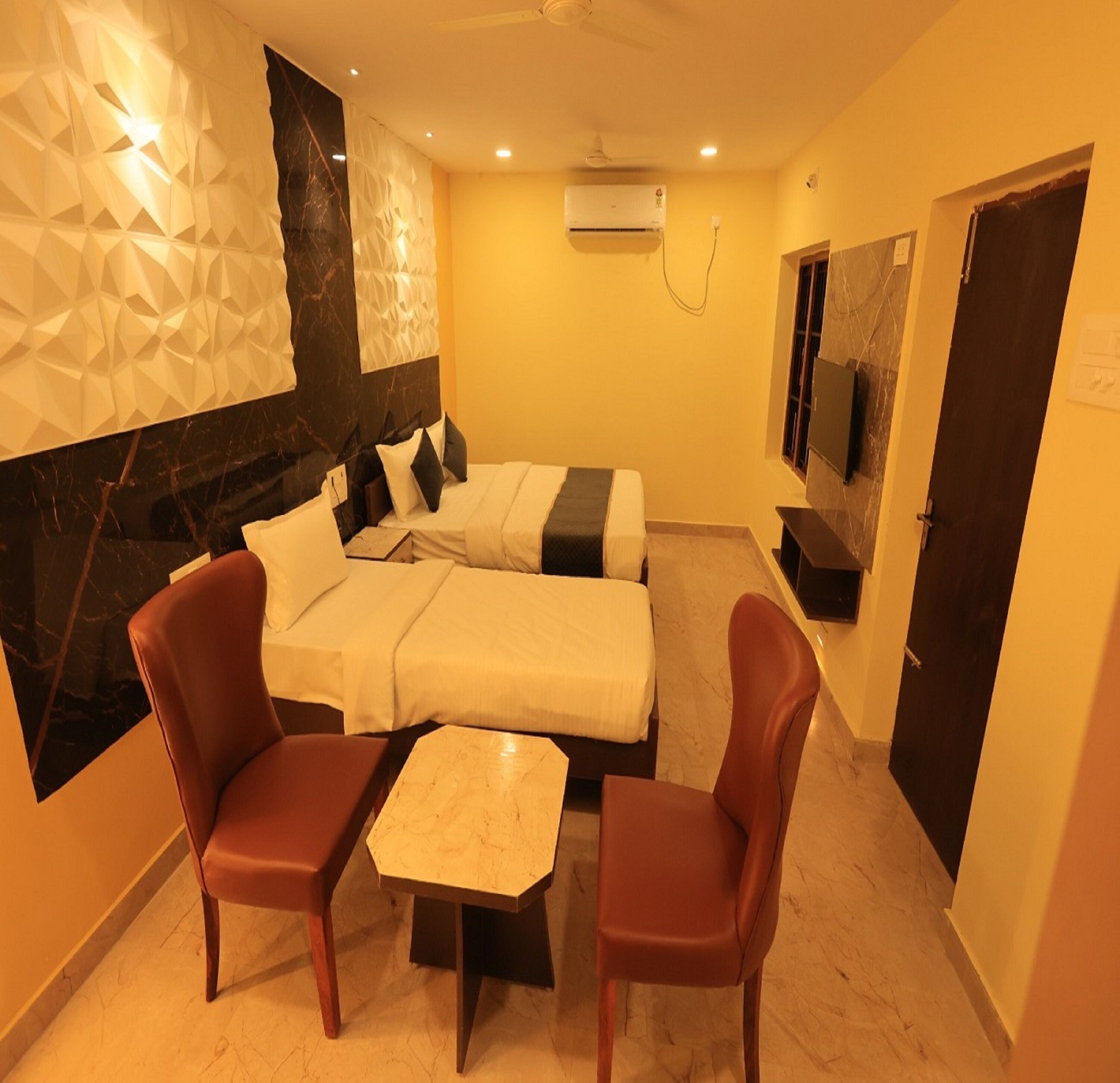 A hotel room with a bed, two chairs, a small table, a marble wall, and an air conditioning unit.
