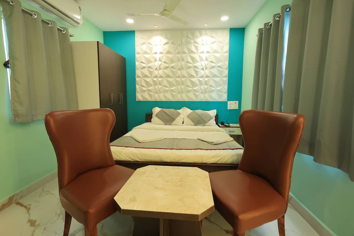 Quarto de hotel com cama, duas cadeiras, uma mesinha, ventoinha no teto e luminária na parede.