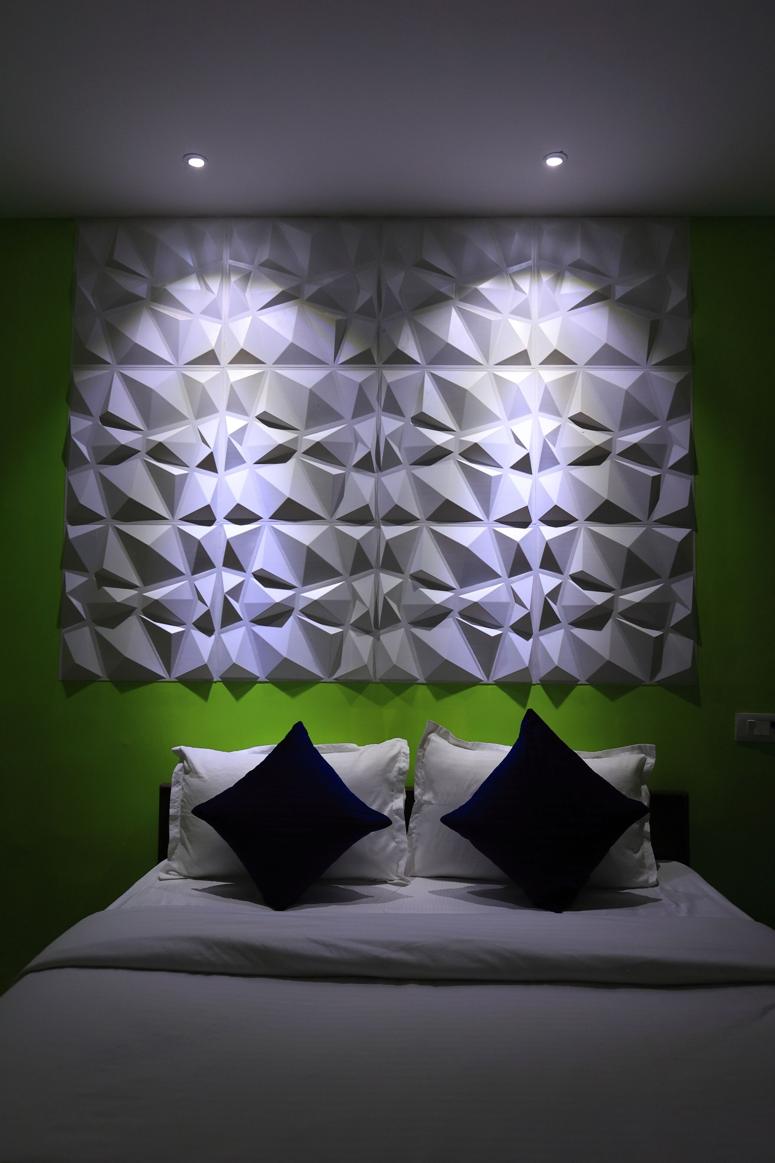 A bed with white bedding and two dark pillows, a geometric wall panel with recessed lighting, and green walls.