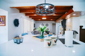 A modern kitchen with white countertops, a hanging light fixture, and a dining area with a sofa and coffee table.