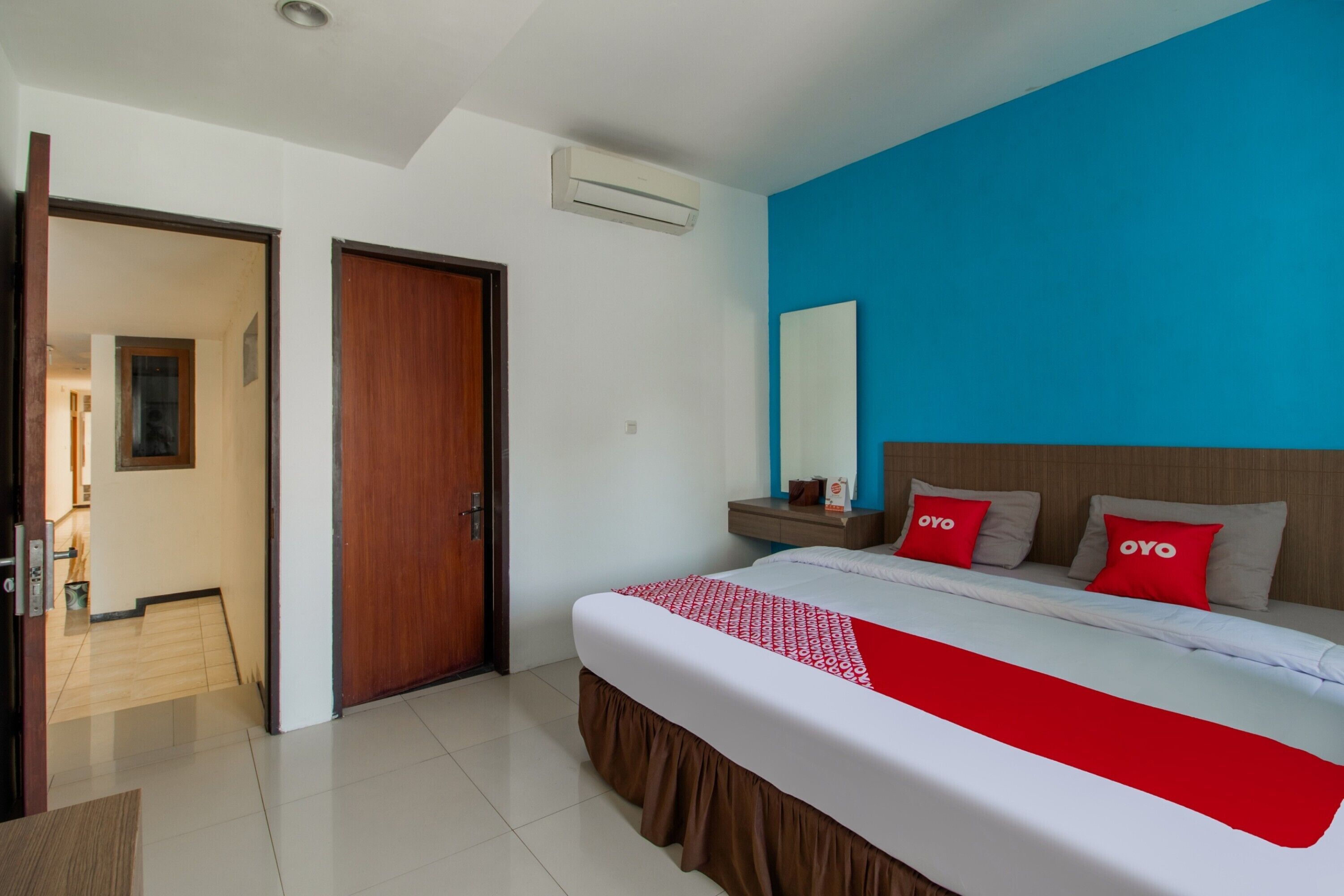 A hotel room with a large bed, a wooden door, a blue accent wall, and a small bedside table with a mirror and a red pillow.