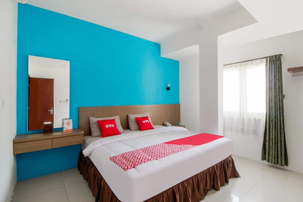 A hotel room with a large bed, a blue accent wall, a wooden nightstand, a mirror, a window with curtains, and a white ceiling.