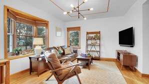 A spacious living room with a large window, a wooden ceiling, a leather sofa, a coffee table, and a flat-screen TV mounted on the wall.