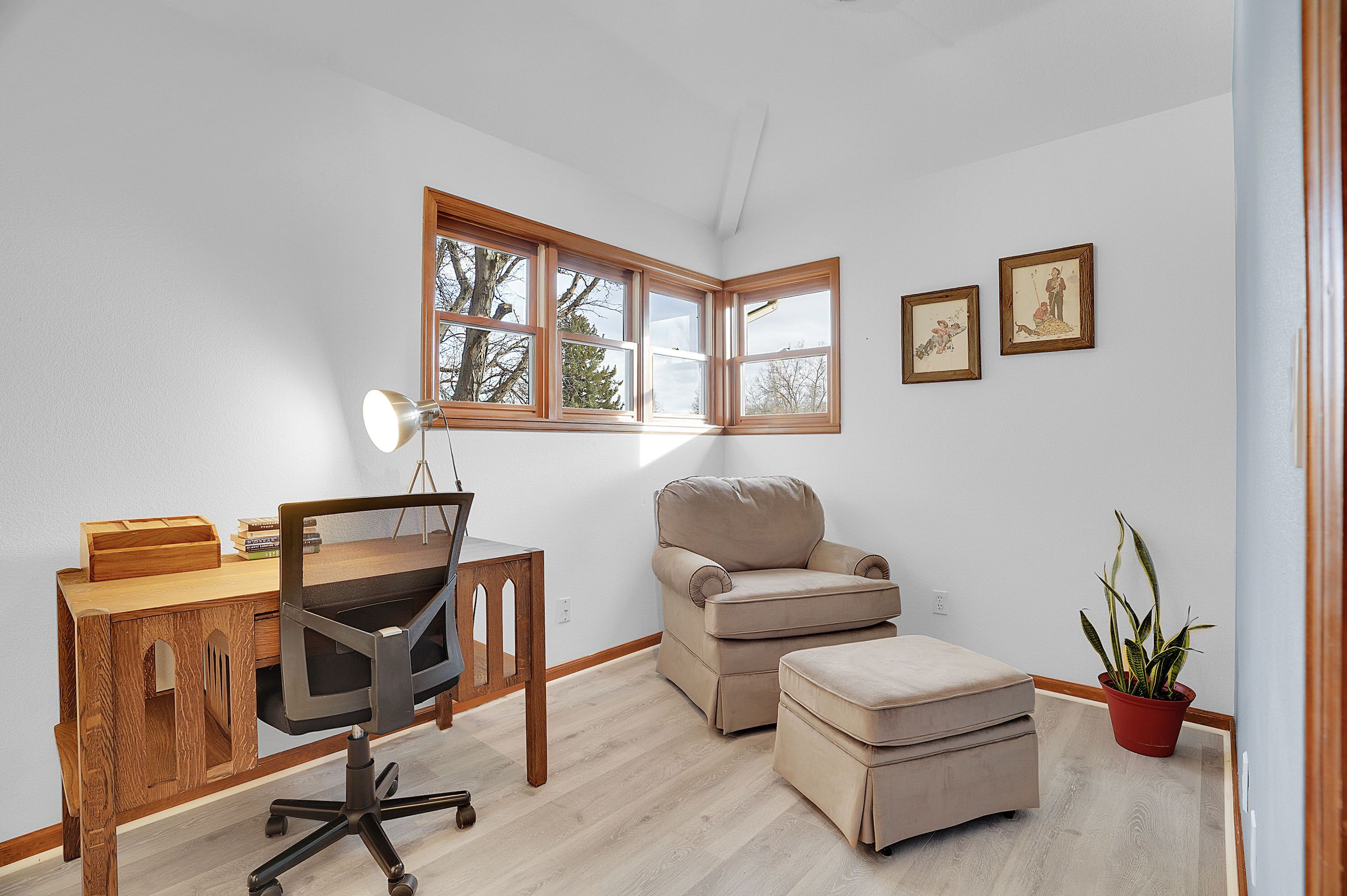 Una habitación con un escritorio de madera, un sillón beigé, un pequeño otoman, una planta en maceta y cuadros en la pared.