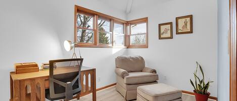 Una habitación con un escritorio de madera, un sillón beigé, un pequeño otoman, una planta en maceta y cuadros en la pared.