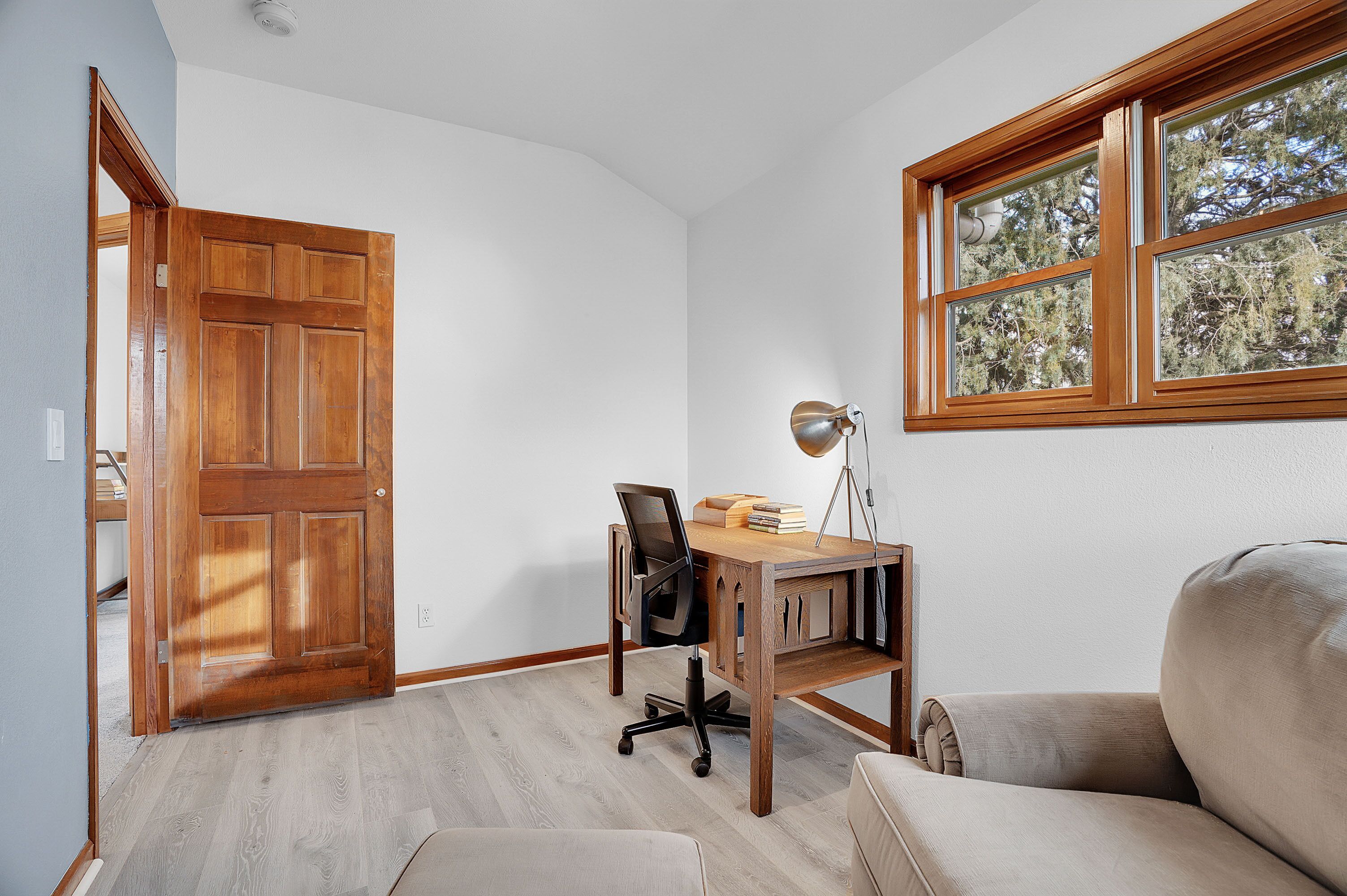A room with a wooden desk, a chair, a lamp, and a window with a view of trees.