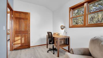 A room with a wooden desk, a chair, a lamp, and a window with a view of trees.