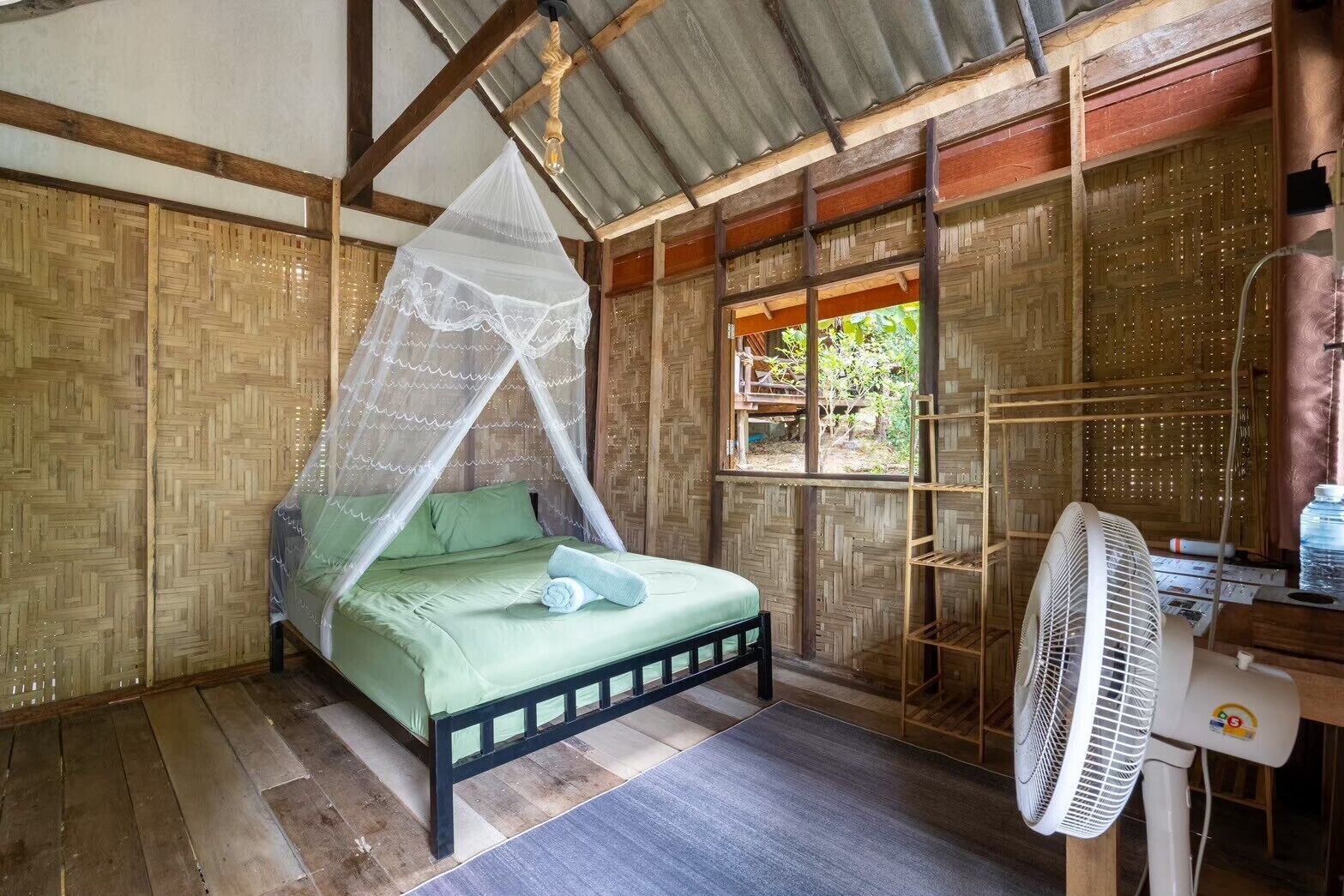 A room with a bed under a mosquito net, a fan, and a window with a view of greenery.