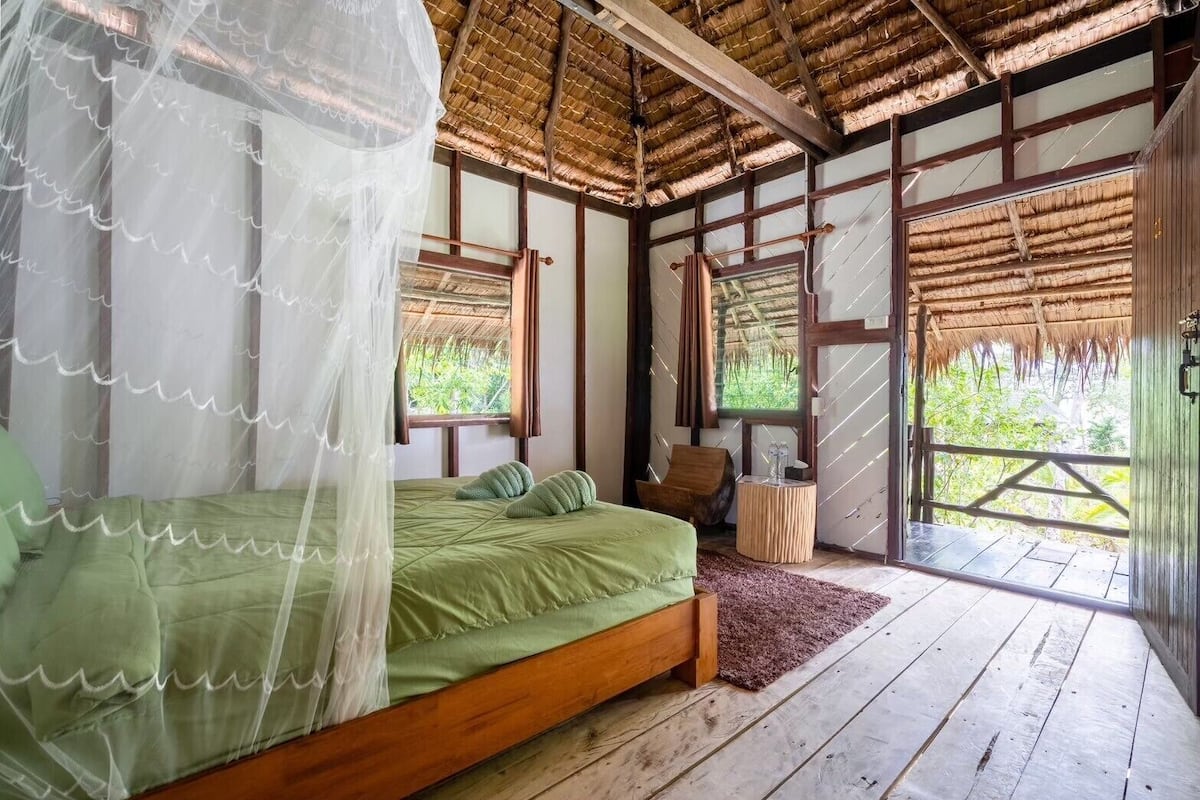A room with a bed, a chair, a small table, and a window with a view of greenery.