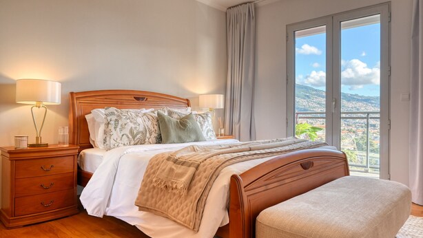 A bedroom with a wooden bed, two bedside tables with lamps, a large window with a view of the city, and a balcony. - Funchal Atrium, a Home in Madeira (Funchal)