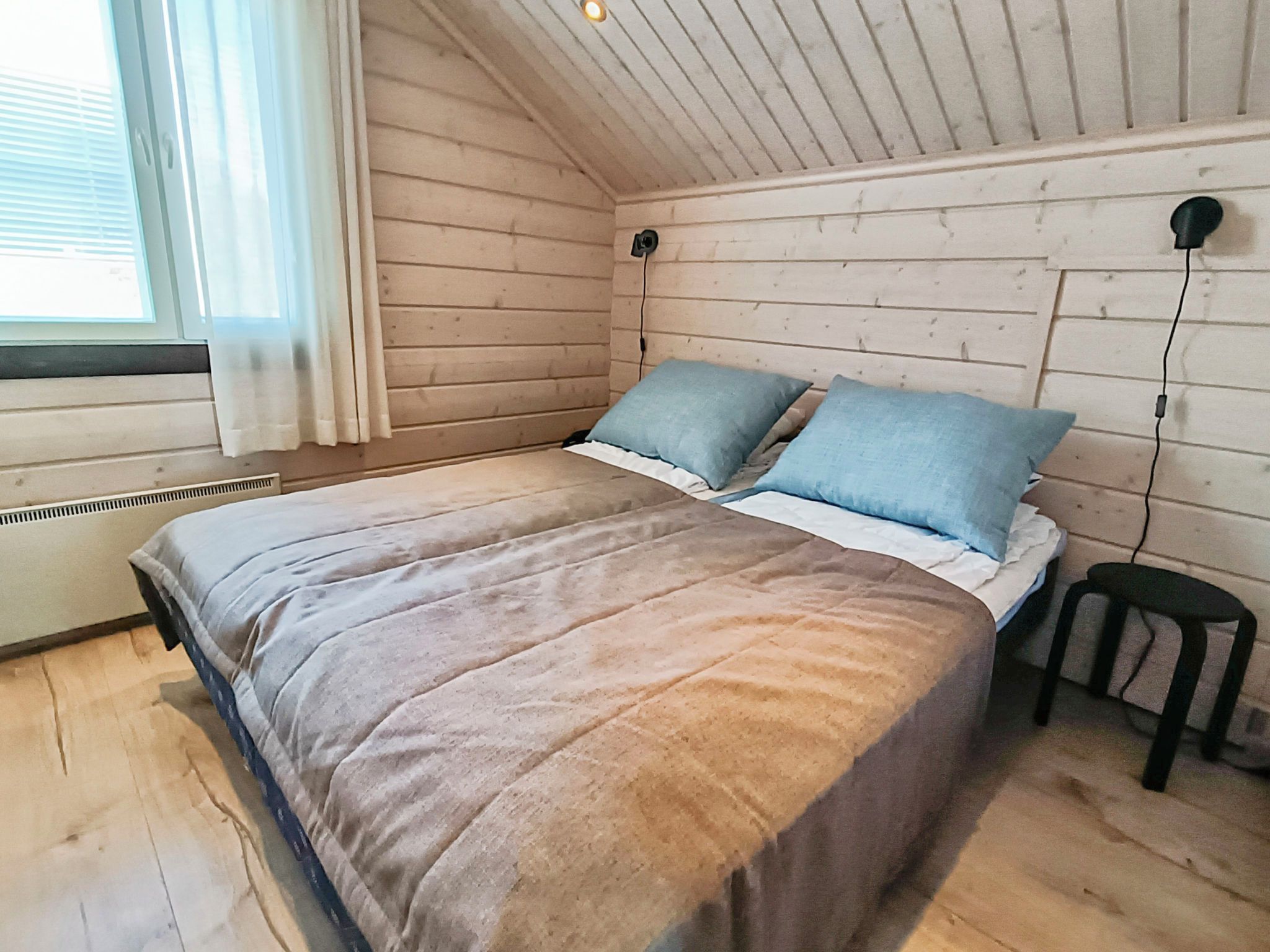 Ein Schlafzimmer mit einem Bett, Kissen, einem Fenster mit Vorhängen, einem Heizkörper und einem Hocker.