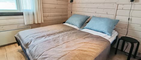 Een slaapkamer met een bed, kussens, een raam met gordijnen, een radiator en een kruk.