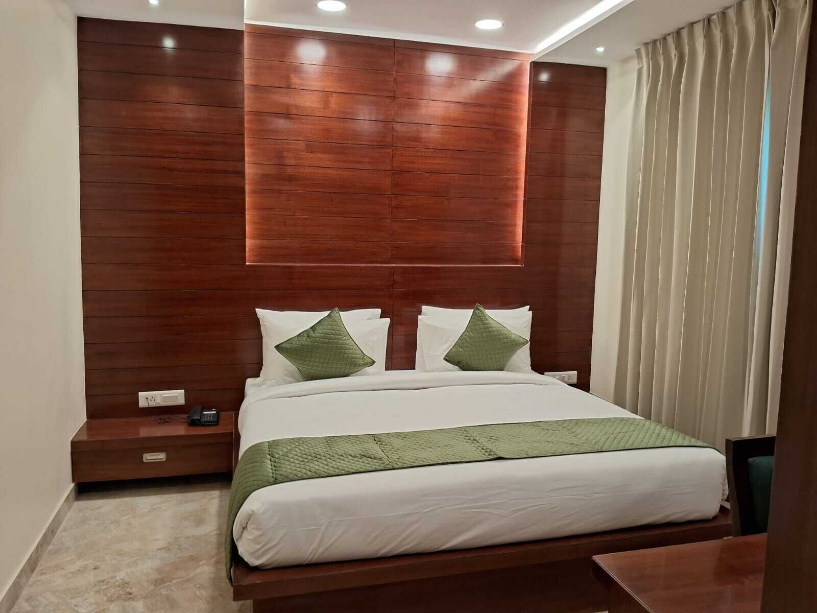 Una habitación de hotel moderna con cabecera de madera, cama con sábanas blancas y almohadas verdes, mesita de noche y cortina en la ventana.