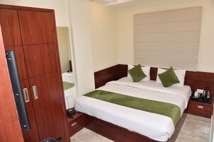 Een hotelkamer met een houten garderobe, een bed met witte beddengoed en groene kussens, een nachtkastje met een lamp, en een raam met jaloezieën.