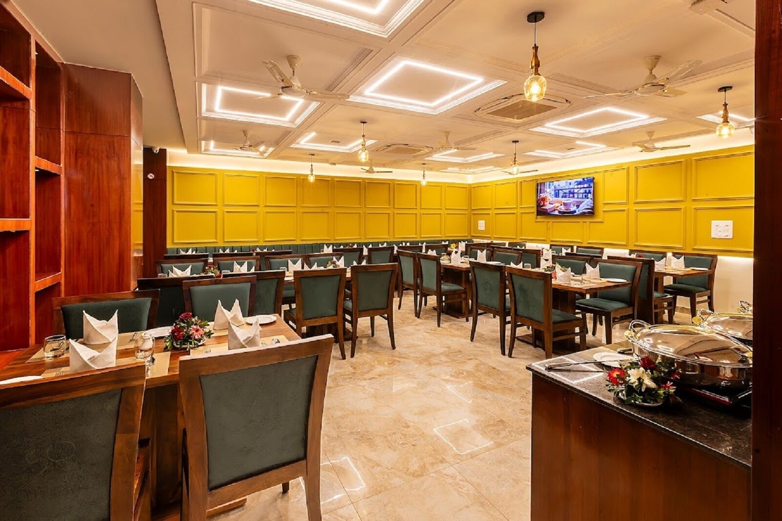 A restaurant with multiple tables and chairs, a television screen on the wall, and ceiling fans.