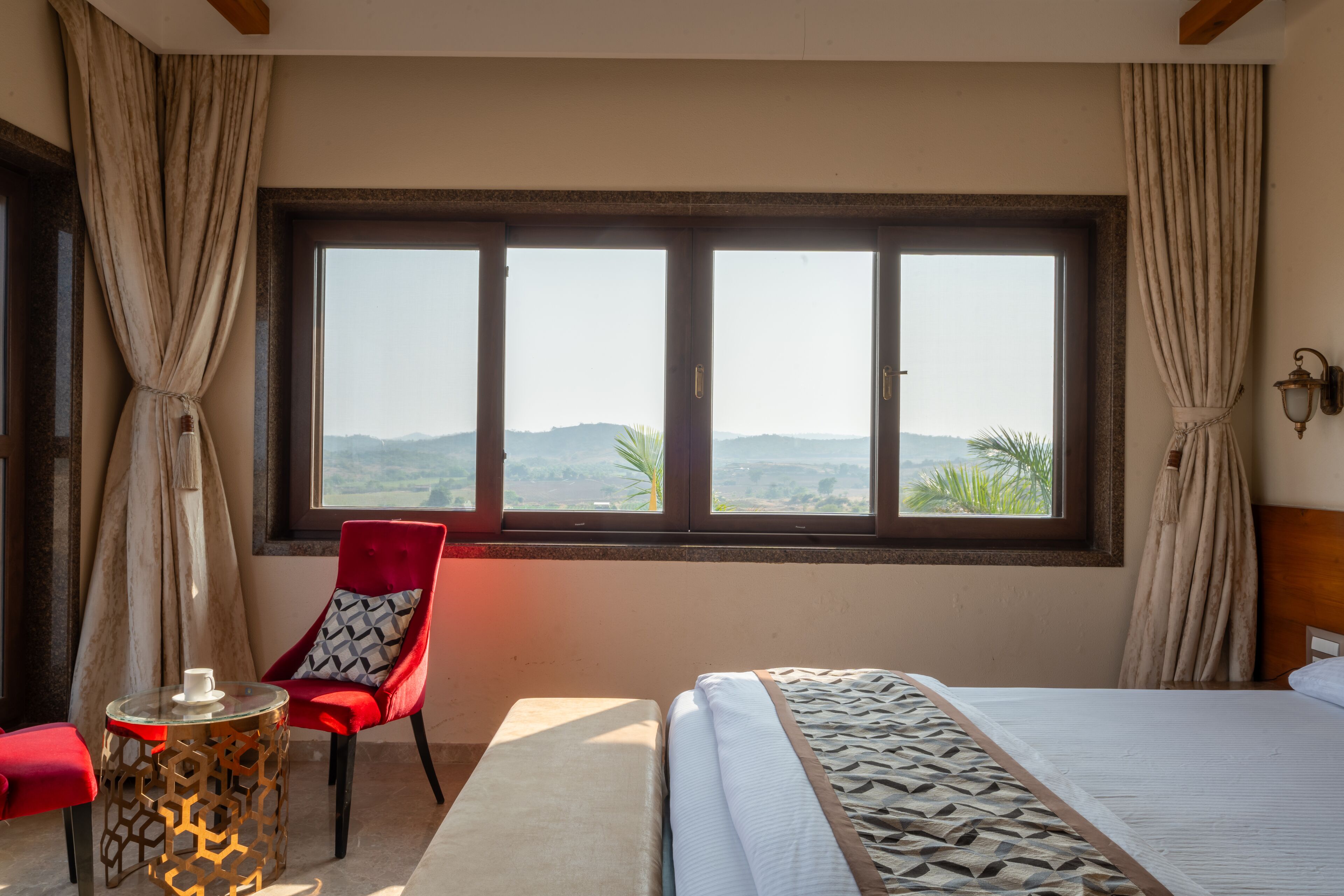 A hotel room with a large window, a bed, a red chair, and a small table with a cup.