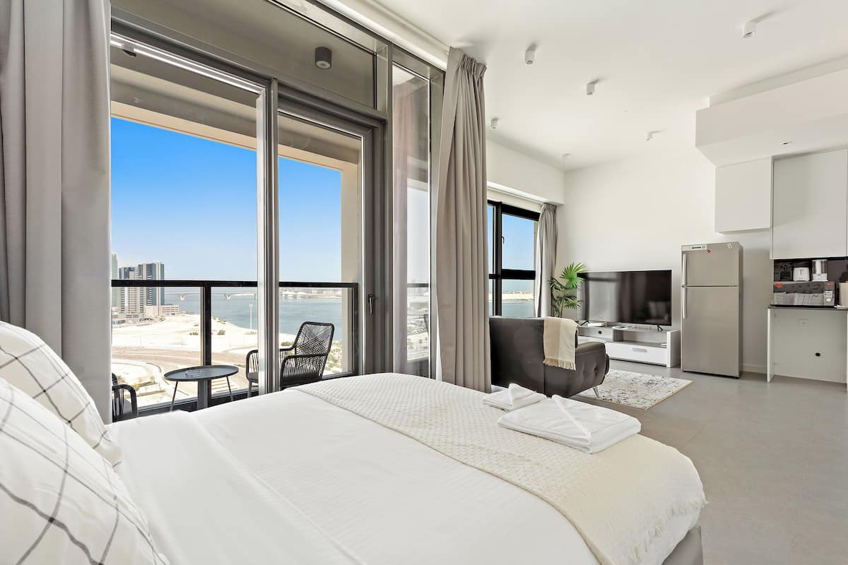 A modern hotel room with a large bed, a balcony with a view of the city and beach, a small dining area, and a kitchenette.