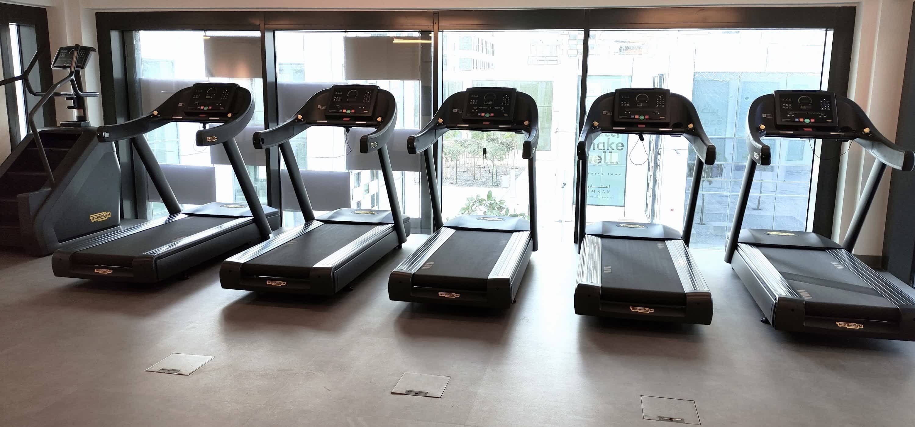 Vier Laufbänder in einem Fitnessstudio mit großen Fenstern und Blick ins Freie.