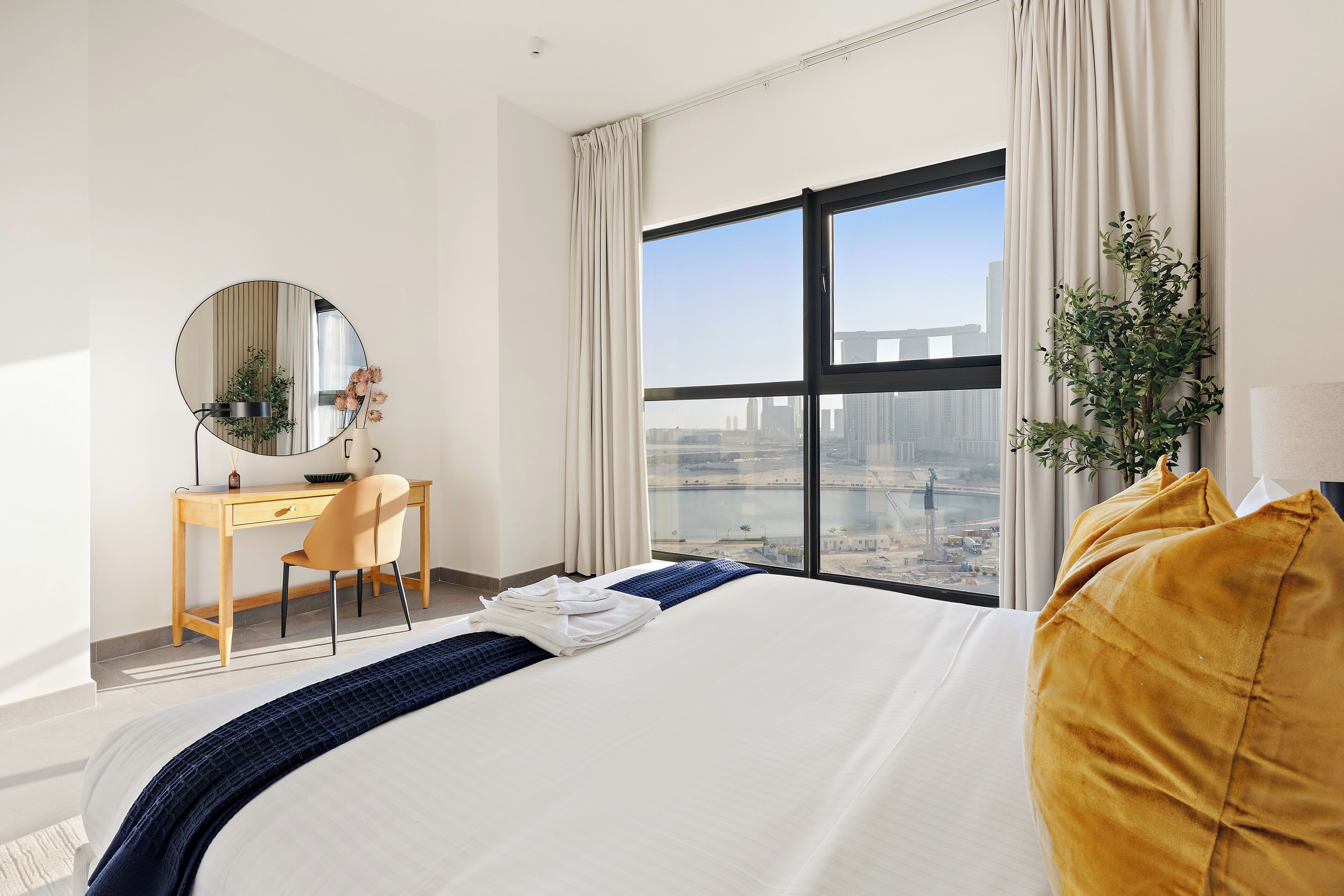 A hotel room with a large bed, a desk with a chair, a round mirror, and a view of the city through a large window.