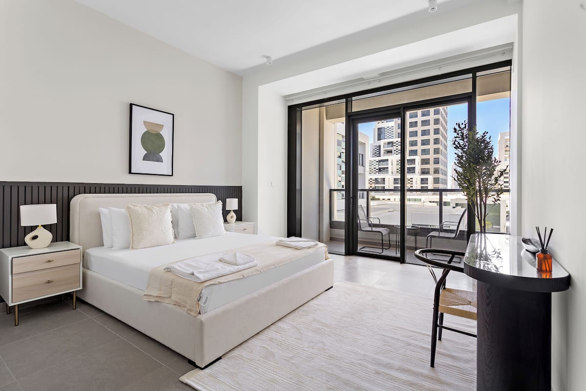 A modern bedroom with a large bed, bedside tables, a desk, and a view of the city through large windows.