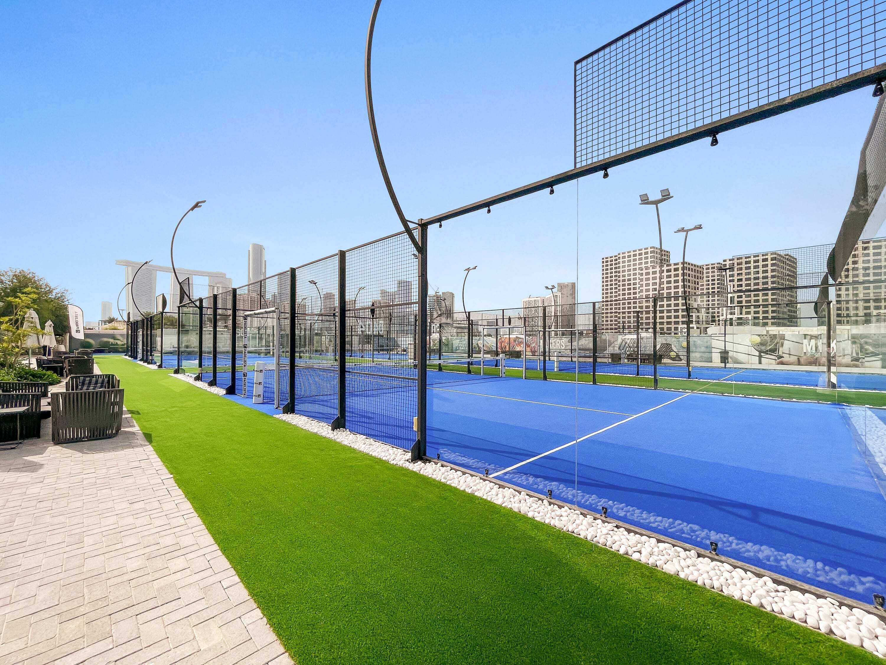 Una cancha de tenis con una red, rodeada por una valla, y un paisaje urbano al fondo.