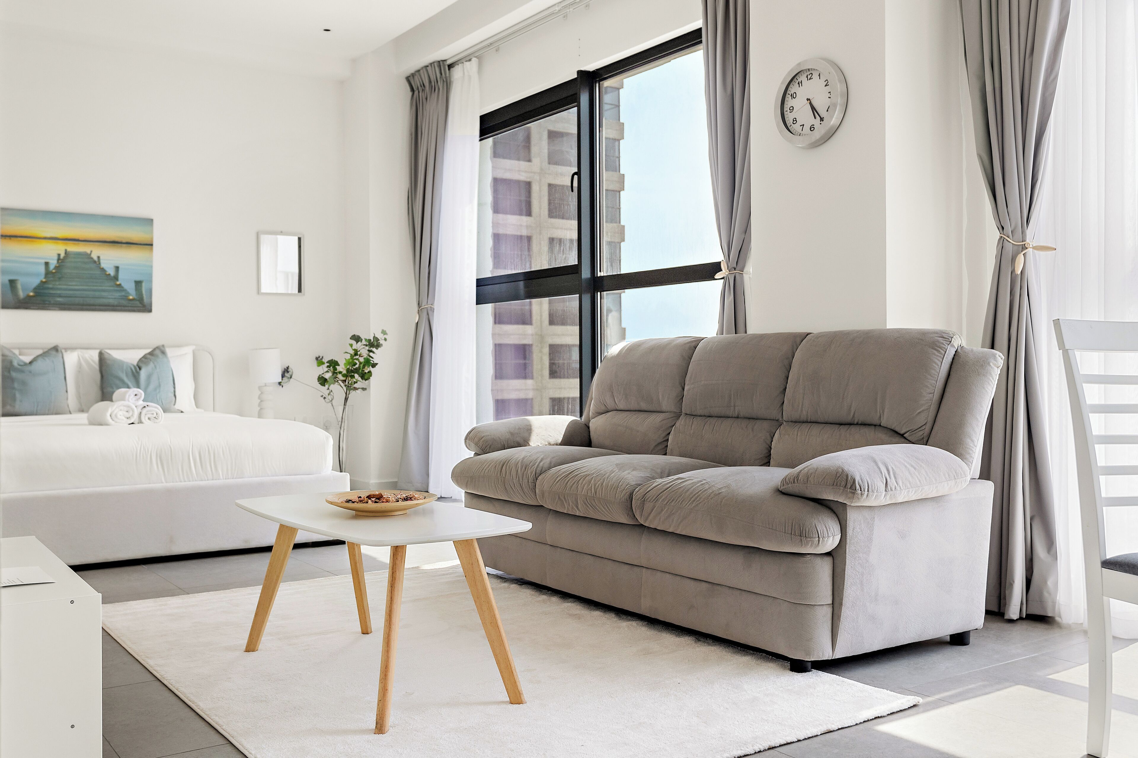 A modern living room with a sofa, a small table, a bed, and a large window with curtains.