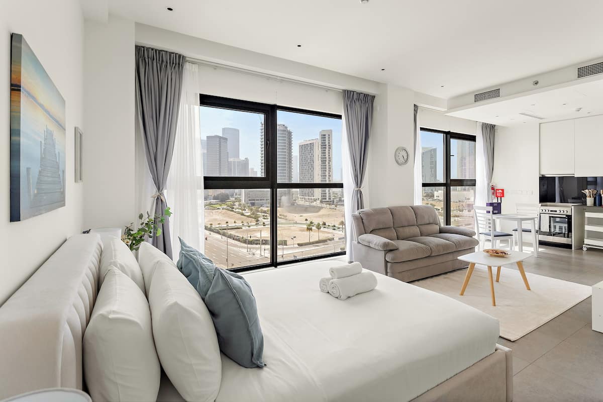 A modern hotel room with a large window, a sofa, a chair, a small table, and a kitchenette.