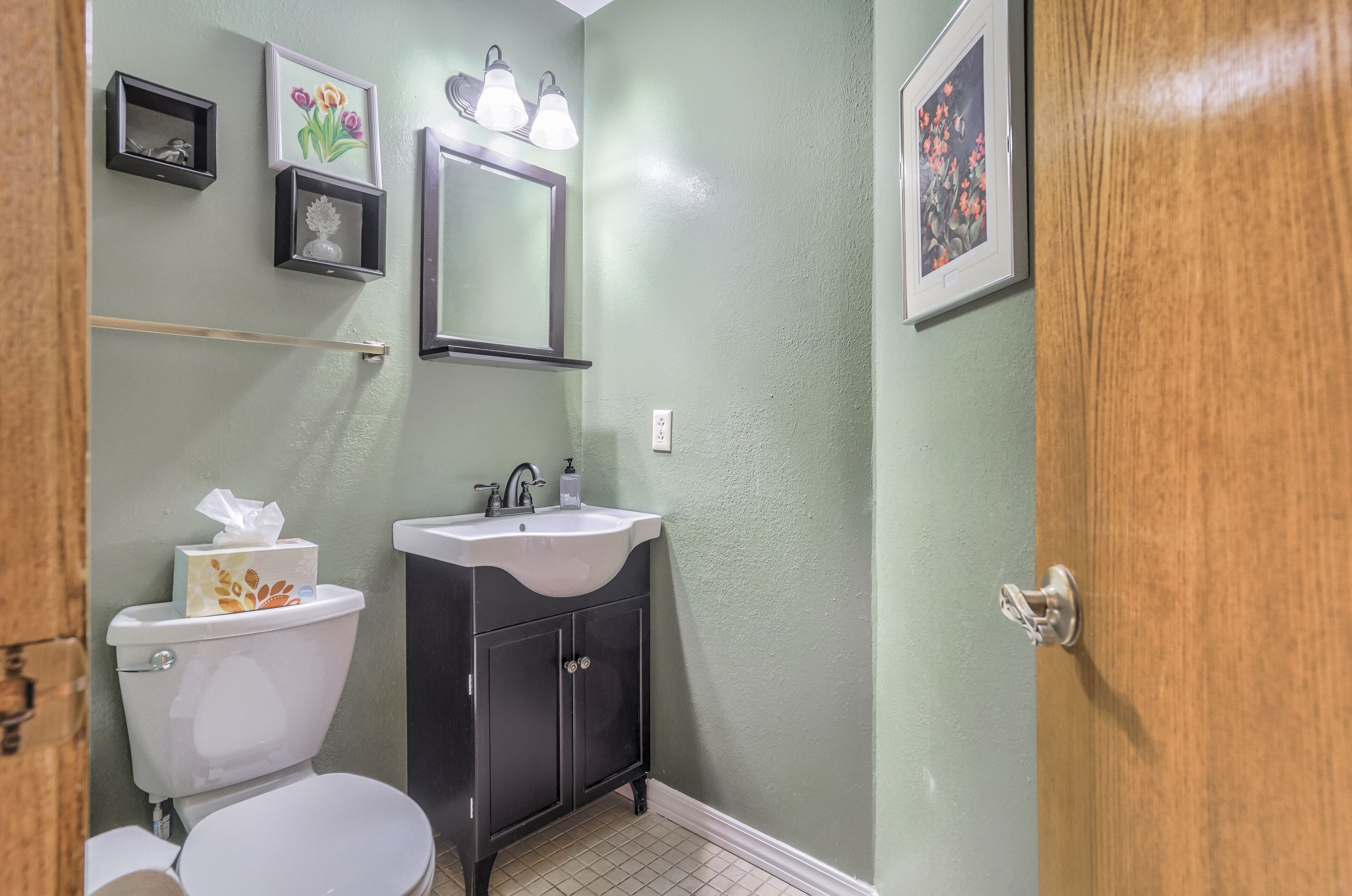 Une salle de bain équipée de toilettes, d’un lavabo et d’un meuble de rangement, agrémentée d’œuvres d’art encadrées et d’un miroir.