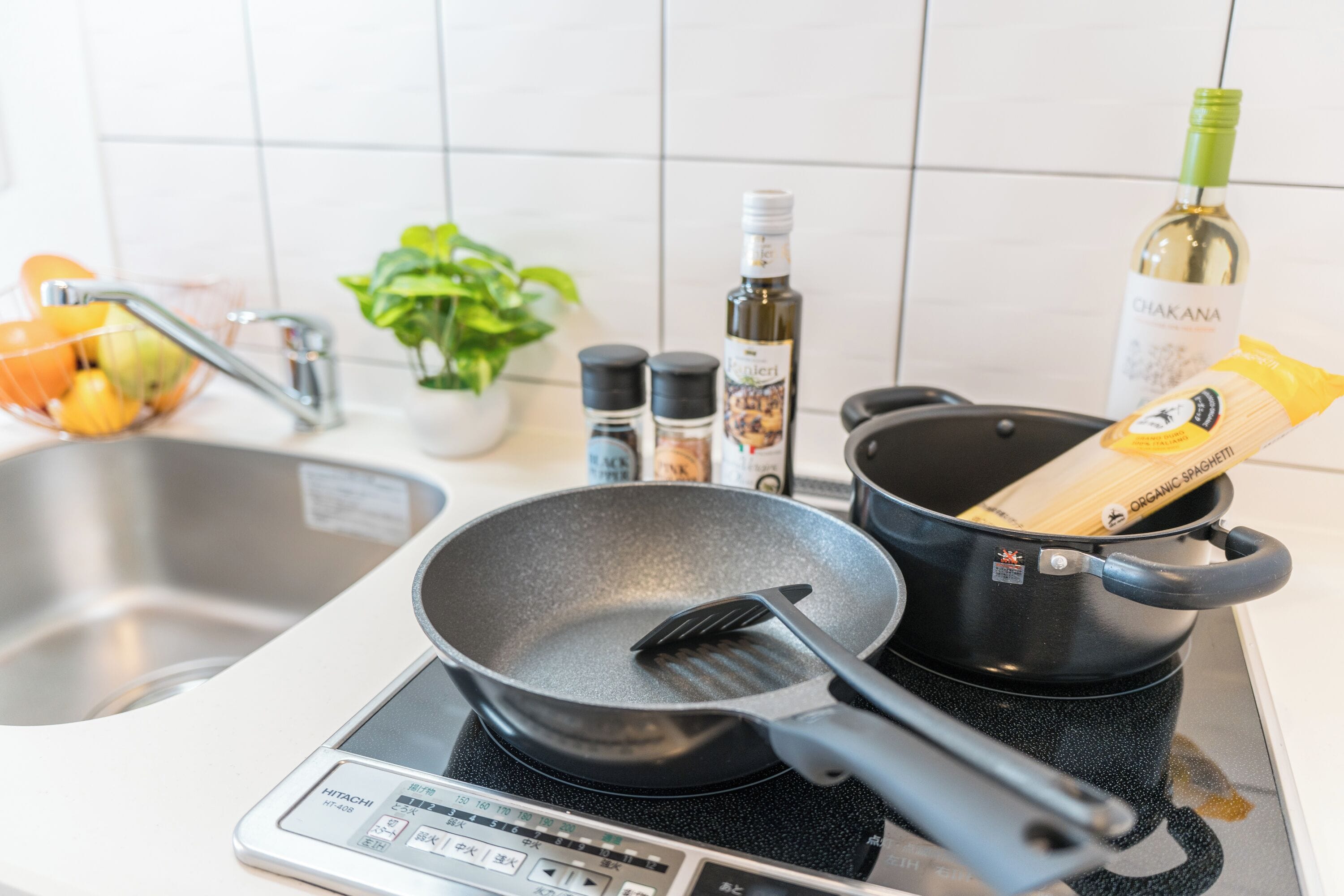 A kitchen with a digital stove, a frying pan, a pot, a bottle of olive oil, a bag of organic spaghetti, and a potted plant.