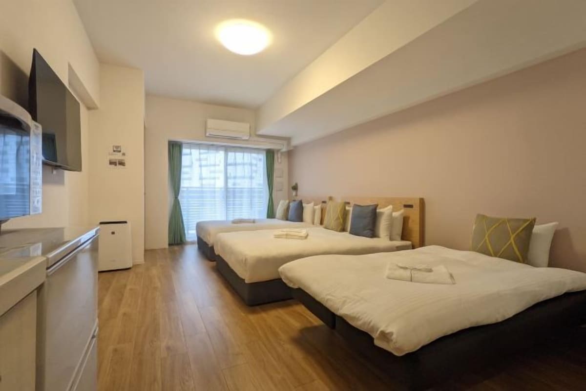 Hotellihuone, jossa on kaksi sänkyä, televisio, pieni keittokomero ja ikkuna, jossa on sälekaihtimet.