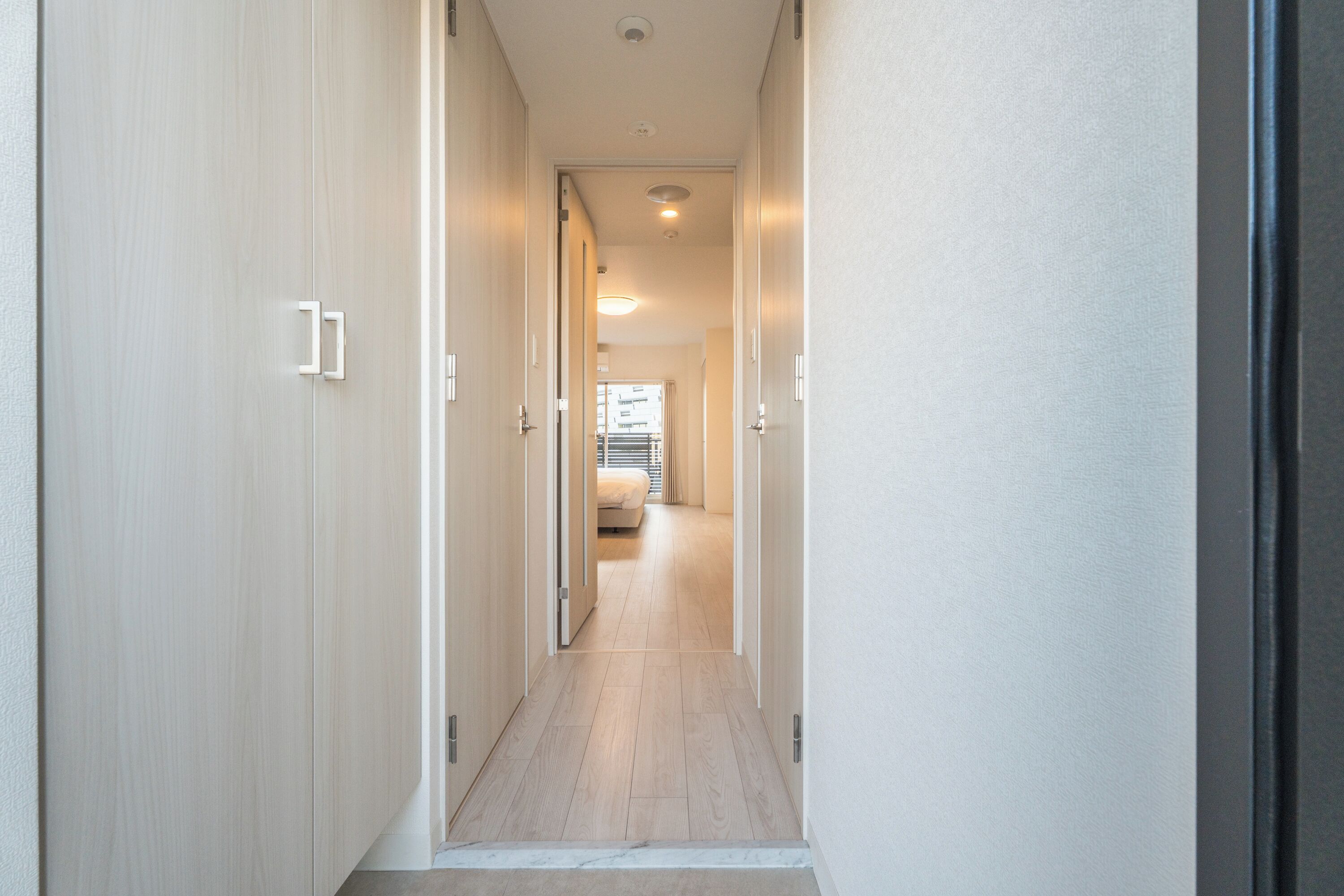 A narrow hallway with wooden flooring leading to a bedroom with a bed and a window.