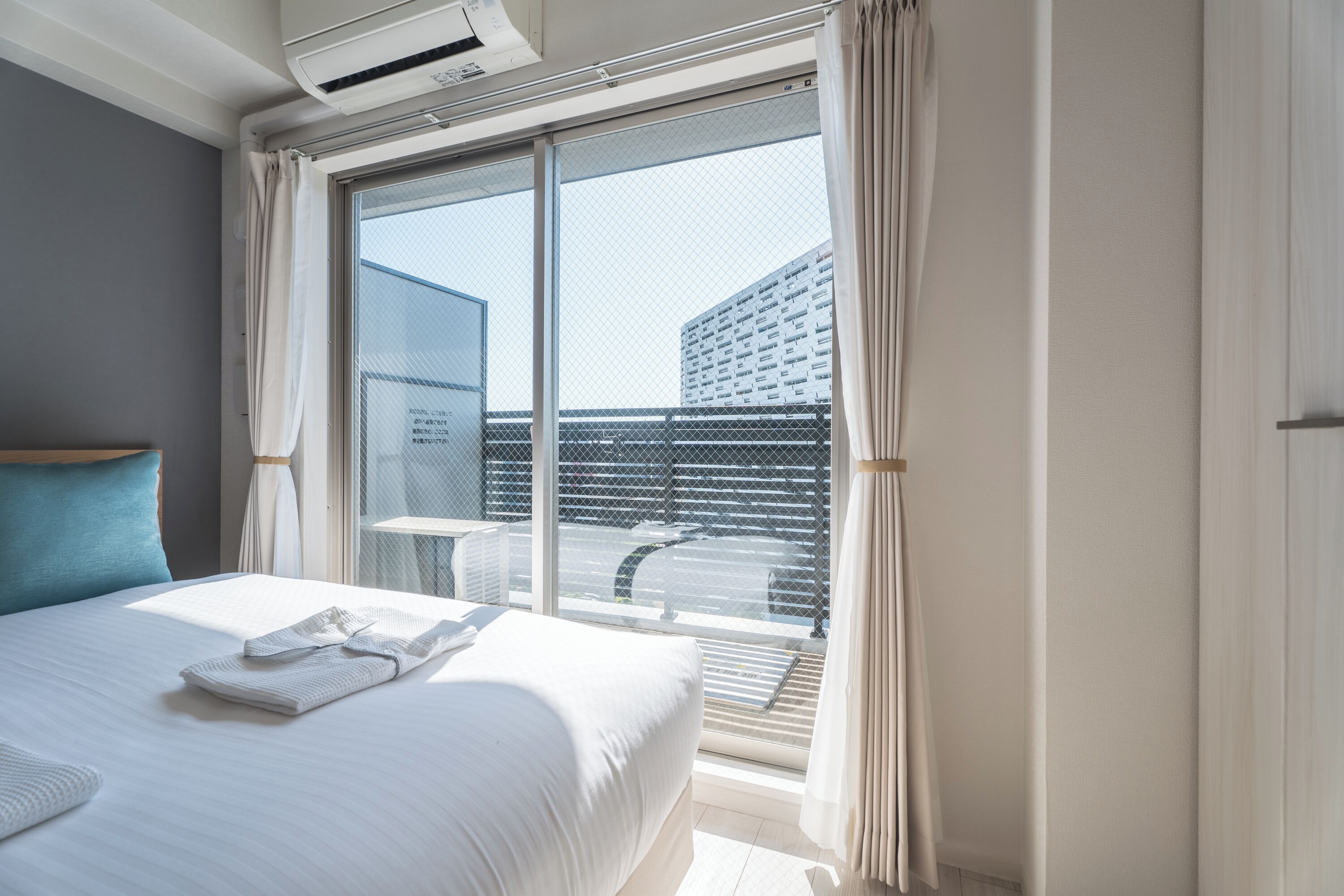 A hotel room with a bed, a window with a view of a building, and a balcony with a railing.