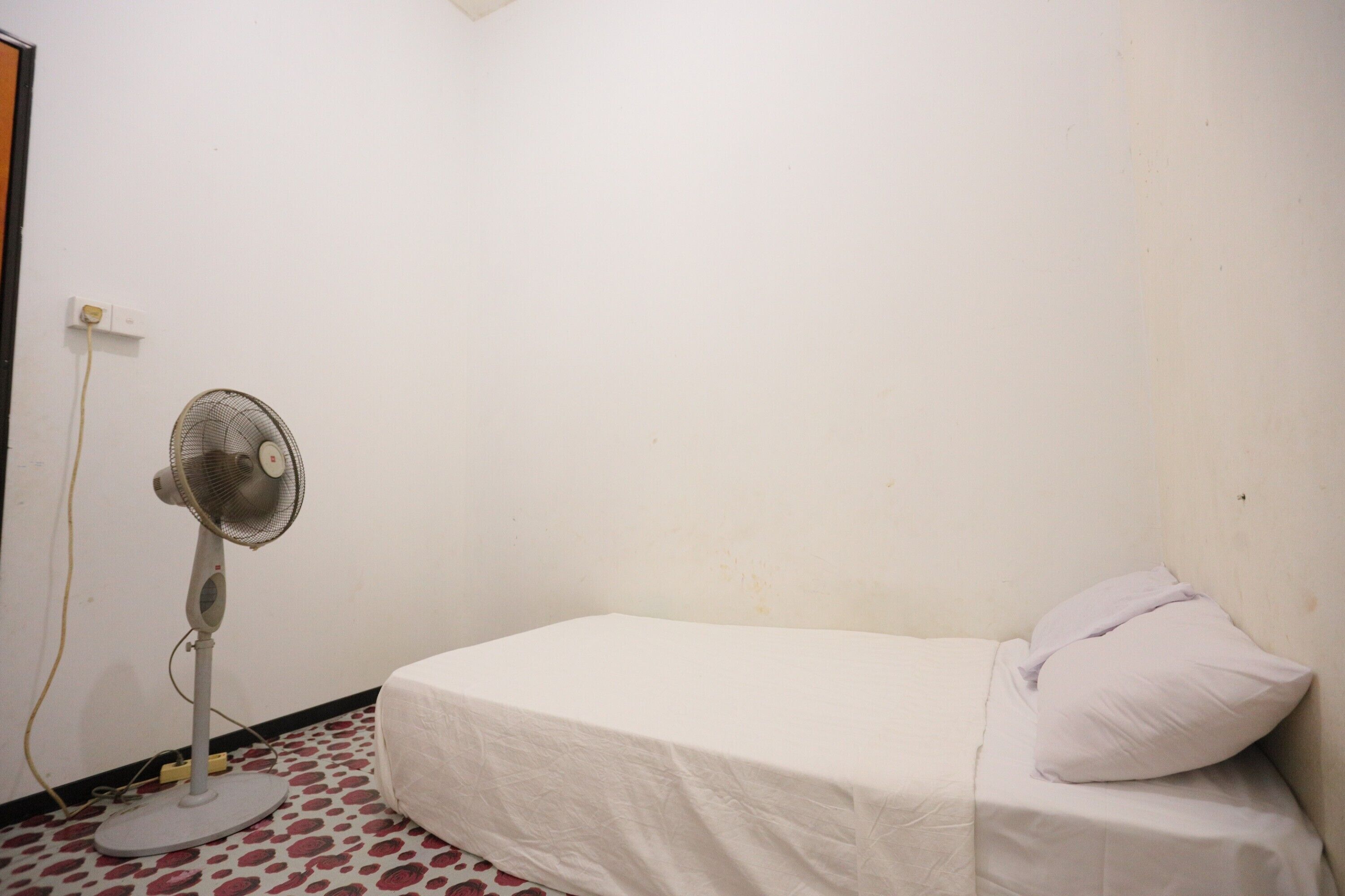 A single bed with a white sheet and a pillow, a standing fan, and a patterned floor.