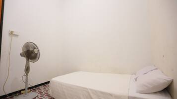 A single bed with a white sheet and a pillow, a standing fan, and a patterned floor.