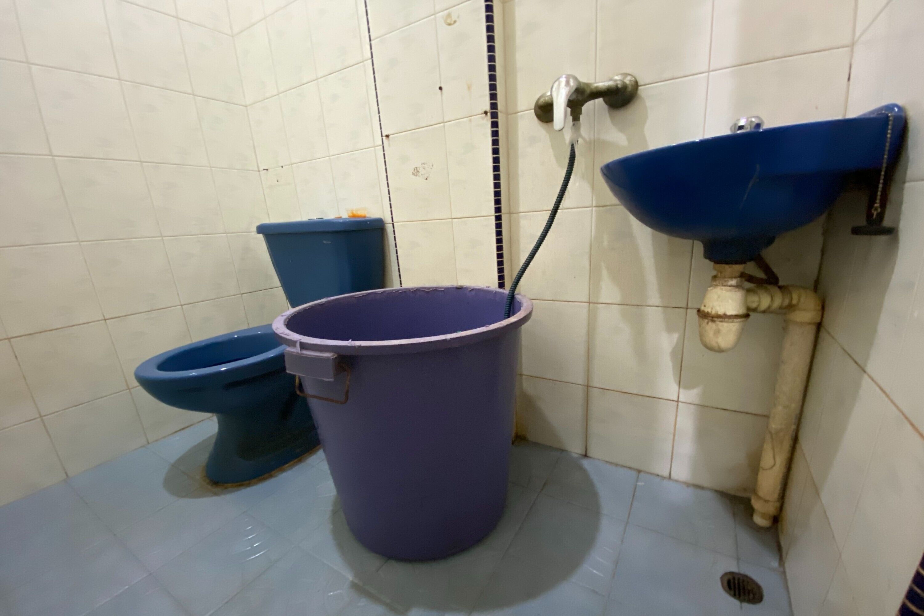 Un baño con dos inodoros, un lavamanos azul y un cubo morado.