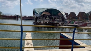 Um barco com área de estar coberta e palco, atracado perto de uma orla com edifícios ao fundo.