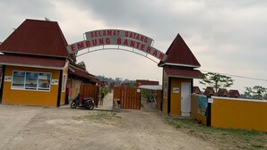 Uma entrada gradeada para uma área residencial chamada Selamat Datang Embing Banteran, com edifícios amarelos e uma estrada de terra.