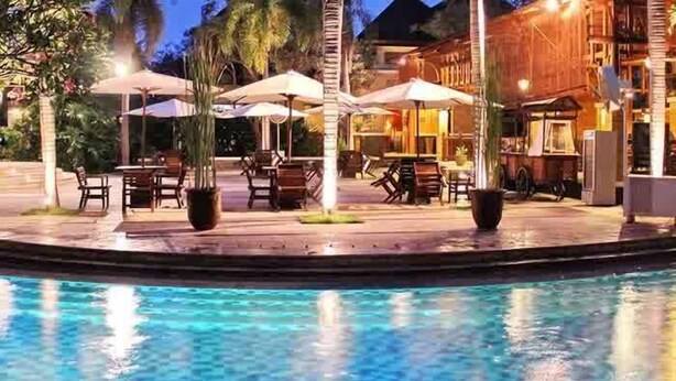 A poolside area with outdoor seating and umbrellas, surrounded by palm trees and a building with a warm facade. - LE DIAN HOTEL & COTTAGES (Serang)